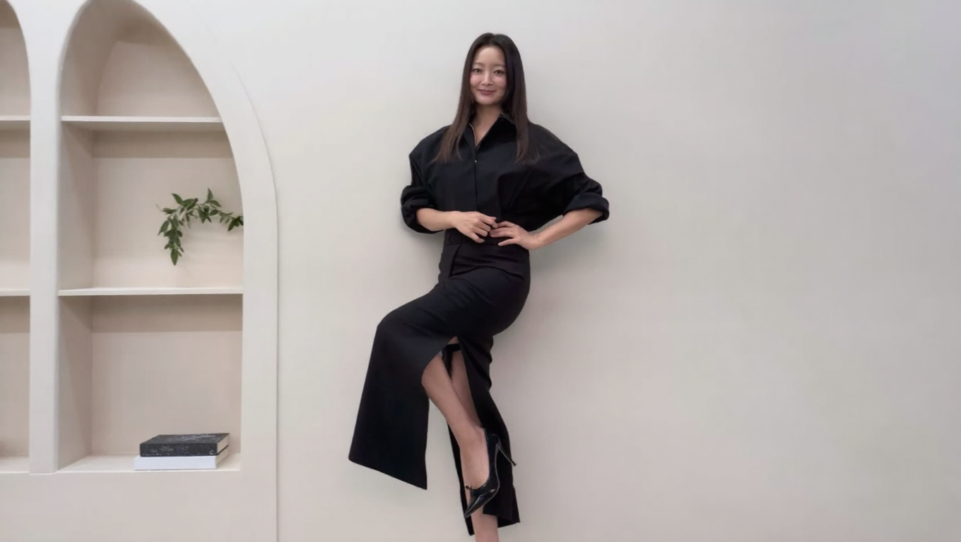 At 48, Kim Hee-sun Fashion proves that a woman's best style decade hasn't started yet. From a Prelin striped polo knit that slims the upper body to a custom butterfly tulle blouse that turns arm coverage into high art, these 15 looks carry one consistent lesson: the right silhouette does more than any diet ever could. If you've already read how Lee Min-jung approaches the same body-proportion challenges, Lee Min-jung Fashion: 15 Slimming Looks is the perfect companion piece to this post. This breakdown covers all 15 looks with the exact styling logic behind each one — plus practical tips you can apply the next time you're standing in front of your wardrobe wondering what to wear. Kim Hee-sun Fashion — The Striped Polo Knit That Slims Without Trying [Photo 1] Kim Hee-sun relaxing in a rattan swing chair, wearing a multi-stripe collar knit top with white bermuda shorts. (Source: Kim Hee-sun's official SNS) Kim Hee-sun Fashion opens with one of the most deceptively simple tricks in the 4060 style playbook. A polo collar creates a clean vertical line from the neck downward, which immediately lengthens the appearance of the neck and jawline. Fine multi-stripe patterns in mixed tones — rather than bold, wide horizontal bands — scatter the eye instead of widening the frame, making the torso read as slimmer overall. The ribbed waist band at the hem gently defines the waistline without any tight tailoring, which is exactly the kind of "invisible shaping" that works best for women in their 40s and 50s. For anyone who finds polo shirts tend to look boxy, try sizing down one size in the body while keeping the shoulder seam in place — the collar will still look relaxed, but the silhouette will read as much sharper. Kim Hee-sun Fashion — White Bermuda Pants That Make Legs Look Longer [Photo 2] Kim Hee-sun standing against a dark wood-panel wall in the same striped polo knit and white wide bermuda shorts. (Source: Kim Hee-sun's official SNS) Kim Hee-sun Fashion uses white bermuda pants here to do something very specific with body proportion. White on the lower half draws the eye downward and outward, which visually extends the leg line beyond where the fabric actually ends. A bermuda length that ends just above or at the knee is the most flattering cut for women in their 40s — it covers the upper thigh and knee area while keeping the lower leg completely visible, which is usually the slimmest part. Pairing a colorful, patterned top with clean white bottoms creates a visual hierarchy that keeps the eye moving upward toward the face, away from any areas around the midsection or hips. If white bermuda shorts feel too casual, a white wide-leg cropped trouser in the same length gives the same leg-lengthening effect with a slightly more elevated feel. Kim Hee-sun Fashion — Adidas Track Pants That Cover Everything Below the Waist [Photo 3] Kim Hee-sun seated on outdoor steps by the water, wearing a black zip-up jacket and Adidas wide-leg track pants with white side stripes. (Source: Kim Hee-sun's official SNS) Kim Hee-sun Fashion proves here that athleisure can be just as body-conscious as any luxury piece. Adidas wide-leg track pants with the signature white side stripe run a vertical line down the full length of the leg, which pulls the eye up and down rather than across — one of the most efficient slimming tricks available in everyday casualwear. The wide leg means the fabric never touches the thigh or calf, so the entire lower body is covered without any clinging or contouring. A grey or dark baseball cap pulled low creates a deliberate air of "not trying," which is a stylistic choice in itself — and it also draws the eye toward the face rather than letting it settle anywhere below the shoulder line. In my opinion, this Adidas track pant silhouette is one of the most underrated options for women in their 40s who want to dress casually without sacrificing proportion control. Kim Hee-sun Fashion — Crop Denim Jacket + Wide Pants for the Perfect Waist Illusion [Photo 4] Kim Hee-sun in front of a dark door, wearing a cropped washed-black denim jacket over a graphic white tee and Adidas wide-leg track pants. (Source: Kim Hee-sun's official SNS) Kim Hee-sun Fashion applies the "short top, long bottom" formula here with precision. A crop jacket that ends at the hip bone creates a visual waistline exactly where it's most flattering — it tells the eye "the legs start here," which makes the lower body appear dramatically longer. Keeping the inner tee long and untucked underneath the jacket means the lower belly is covered even when the jacket is open, which solves one of the most common concerns for women in their 40s without requiring any tight layering. The contrast between the heavyweight washed denim jacket and the fluid wide-leg track pants is also a textural balance — structured top, soft bottom — that keeps the overall look from looking too casual or too stiff. Practical tip: if you don't want to invest in a designer crop jacket, a thrifted or vintage oversized denim jacket worn with the sleeves rolled once and the body left fully unbuttoned reads almost identically from a distance. Kim Hee-sun Fashion — Sweatshirt + Adidas Maxi Skirt for Unexpected Elegance [Photo 5] Kim Hee-sun standing in front of a pink-winged street mural, wearing a light grey sweatshirt and a navy Adidas maxi skirt with side stripe detail. (Source: Kim Hee-sun's official SNS) Kim Hee-sun Fashion takes a sharp turn here — and it works entirely because of length. Pairing a relaxed sweatshirt with a floor-length skirt transforms both pieces: the sweatshirt reads as intentional rather than lazy, and the maxi skirt elevates what would otherwise be a purely casual sportswear item. The Adidas side stripe on a maxi skirt length draws one unbroken vertical line from hip to floor, which is as close to an optical height-booster as casualwear gets. For women in their 40s and 50s who are self-conscious about thighs, calves, or ankles, a maxi-length athletic skirt is genuinely one of the best-kept secrets in everyday dressing — full coverage, zero effort, and the vertical stripe does the rest. A similar look is achievable with any wide, floor-length skirt in a dark solid color combined with a relaxed-fit sweatshirt in a neutral tone. Kim Hee-sun Fashion — Butterfly Tulle Blouse That Covers Arms Like Nothing Else [Photo 6] Kim Hee-sun backstage, wearing a custom white butterfly-embroidered tulle blouse with voluminous puff sleeves and ruffled details. (Source: Kim Hee-sun's official SNS) Kim Hee-sun Fashion reaches its most dramatic moment here — and the arm-coverage lesson is unmissable. Voluminous tulle sleeves that gather from shoulder to wrist cover the upper arm completely while the sheer fabric means the arm isn't hidden so much as softened — it becomes a gentle shape rather than a defined silhouette. The butterfly and floral embroidery scattered across the sleeve surface draws the eye to the decorative detail rather than the arm underneath, which is an advanced version of the "distraction with beautiful things" approach to body coverage. For women who avoid sleeveless or short-sleeved tops because of upper-arm concerns, a voluminous tulle or organza sleeve is the most elegant solution — it adds volume to the sleeve that reads as artistic rather than compensatory. The high mock-neck collar in the same sheer fabric is also doing quiet work on neck coverage, hiding any neck area concerns without the stiffness of a traditional turtleneck. Kim Hee-sun Fashion — Mirror Selfie That Shows the Full Tulle Look [Photo 7] Kim Hee-sun taking a mirror selfie in the same white butterfly tulle blouse, showing the full upper-body silhouette. (Source: Kim Hee-sun's official SNS) Kim Hee-sun Fashion gives us the full picture here from a different angle, and the waist detail becomes clear. The white tulle blouse tapers through the torso, so despite the enormous volume of the sleeves and collar, the body itself reads as slim — this is the classic "balloon sleeve, fitted body" approach that works so reliably for mature women's styling. Pulling the hair back into a simple updo fully reveals the high neckline detail and lets the embroidered collar speak without competition. The mirror-selfie angle at approximately 45 degrees is also the most flattering shooting angle for a puff-sleeve top — it shows the volume of one sleeve while keeping the face and torso clearly visible. Kim Hee-sun Fashion — Backstage Tulle Look: Why Sheer Is More Slimming Than Solid [Photo 8] Kim Hee-sun backstage in the same white tulle blouse, hands raised to her cheeks, surrounded by the costume rack. (Source: Kim Hee-sun's official SNS) Kim Hee-sun Fashion gives us one more view of this extraordinary blouse, and the styling principle becomes even clearer. Sheer fabric over a solid inner base creates a layered visual depth that solid fabric can't — the eye perceives the arm or shoulder as "existing inside something beautiful" rather than "being covered up." For women in their 40s and 50s who have been avoiding sheer fabrics because they feel too revealing, the key is the inner base: a skin-tone or white slip underneath a white sheer outer means full modesty with all the visual lightness of sheer fabric. The ruffled collar gathered around the neck and shoulders in this look also does the additional work of widening the appearance of the shoulders, which naturally makes the face look smaller and the overall silhouette more balanced. Kim Hee-sun Fashion — Scallop-Trim Blouse That Frames the Face Perfectly [Photo 9] Kim Hee-sun wearing a white organza blouse with black scallop-edge trim and bold striped collar, adjusting her hair. (Source: Kim Hee-sun's official SNS) Kim Hee-sun Fashion pivots to a completely different silhouette here — structured and graphic rather than dreamy and soft. A wide flat collar with bold black scallop-edge trim creates a strong horizontal frame around the collarbone area, which draws the eye upward and keeps attention on the face and neck. Puff sleeves that gather gently at the wrist add feminine volume through the arm without any tight cling — the same sleeve construction used in the tulle blouse, here executed in a crisper organza. The gold statement earring visible on one side adds a warm, high-contrast accent that breaks the black-and-white graphic palette and prevents the look from reading as too severe for daytime wear. Kim Hee-sun Fashion — Black Ribbed Dress with White Bow That Redefines Monochrome [Photo 10] Kim Hee-sun in profile beside a clothing rack, wearing a structured black ribbed outfit with a large white bow at the neckline. (Source: Kim Hee-sun's official SNS) Kim Hee-sun Fashion uses one of the oldest tricks in the book here — a large white accent placed directly at the neckline — and proves it still works at the highest level. A single white bow or contrast collar at the neck creates an instant focal point that pulls every eye upward toward the face, which is exactly where you want attention in a monochrome look. The ribbed black base underneath creates vertical texture that elongates the entire silhouette, while the structured sleeves with contrast trim keep the arms looking defined rather than soft. This is a look that works because it commits fully — the bow is large enough to be intentional, the black is deep enough to anchor it, and neither element apologizes for itself. Kim Hee-sun Fashion — All-Black Shirt Dress That Lengthens the Entire Body [Photo 11] Kim Hee-sun standing in a room interior, wearing a full all-black oversized shirt tucked into a black H-line midi skirt, pink watch accent. (Source: Kim Hee-sun's official SNS) Kim Hee-sun Fashion shows the power of monochrome dressing most clearly in this look. When the top and bottom are the same color, the eye reads the body as one continuous unbroken shape — which is significantly taller and slimmer-looking than any two-color combination could achieve. A single pop of color at the wrist — here, a pink watch — gives the eye somewhere to rest without breaking the vertical line of the all-black silhouette. Tucking a voluminous shirt into a high-waisted skirt raises the visual waistline to its most flattering position and keeps the shirt from adding bulk around the midsection. For women in their 40s and 50s who feel self-conscious about the abdominal area, a half-tuck (front only, sides and back left loose) achieves the same waistline effect with more coverage over the lower belly. Kim Hee-sun Fashion — Black Side-Slit Skirt That Shows Less but Looks Like More [Photo 12] Kim Hee-sun posing against a white curved wall, wearing an oversized black shirt with a black H-line midi skirt featuring a deep side slit and black pointed heels. (Source: Kim Hee-sun's official SNS) Kim Hee-sun Fashion delivers one of its most powerful body-proportion lessons here through a single design detail: the side slit. A floor-length or midi skirt that covers the entire leg creates a sleek, elegant silhouette — but a deep side slit that opens with each step reveals only the lower shin and ankle, which are typically the slimmest parts of the leg. This creates the impression of leg length and confidence without the exposure of a mini or thigh-high slit, which is exactly the balance most women in their 40s are looking for. Black pointed-toe heels elongate the foot and ankle line below the slit, adding a final visual extension that makes the leg appear to continue well below the hemline. Kim Hee-sun Fashion — Side Profile in All-Black That Shows the Real Silhouette Power [Photo 13] Kim Hee-sun from the side against the same curved white wall, arms crossed, the full length of the black skirt and slit visible. (Source: Kim Hee-sun's official SNS) Kim Hee-sun Fashion gives us the side view here, and the full silhouette impact of the H-line skirt becomes unmistakable. A true H-line skirt — straight from hip to hem with no flare or taper — creates the most elongating silhouette of any skirt shape for women with fuller hips or thighs, because the fabric never clings at the widest point. Crossing the arms at the front naturally draws the fabric of the loose shirt inward at the center, creating the appearance of a defined waistline without any belt or tight tailoring. The overall effect — side-parted straight hair, long black column silhouette, single slit opening — is the closest thing to a universally flattering formula that exists in women's fashion. Kim Hee-sun Fashion — Lemon Yellow Shirt + Black Knitwear for the Brightest Face Trick [Photo 14] Kim Hee-sun holding a birthday cake with a lit candle, wearing a lemon-yellow cotton shirt with a black knit cardigan draped over the shoulders. (Source: Kim Hee-sun's official SNS) Kim Hee-sun Fashion saves its most practical everyday lesson for Look 14. A pale lemon-yellow shirt placed directly at face level acts as a natural reflector — it bounces warm, golden light upward onto the skin, which instantly makes the face look brighter and more rested. Draping a dark cardigan over the shoulders rather than wearing it properly is one of the most effective tricks for mature dressing: it adds a layer of coverage to the shoulder and upper back area while keeping the look deliberately casual rather than bundled up. The black cardigan draped over yellow also creates a strong color contrast that keeps the eye moving between the two tones, preventing any single area of the body from becoming the visual focus. For women who want to try this "draped knit" technique, the key is letting the cardigan sit toward the back of the shoulders rather than the front — this keeps the chest area open and avoids adding bulk at the front of the torso. Kim Hee-sun Fashion — Birthday Look That Proves Casual Is the New Elegant at 48 [Photo 15] Kim Hee-sun seated at a table with a birthday cake, wearing the same lemon yellow shirt and black draped cardigan, hair loose, smiling softly. (Source: Kim Hee-sun's official SNS) Kim Hee-sun Fashion closes on its most human note — a birthday, a cake, and a look that asks nothing of the viewer except to notice how completely at ease she is. The lemon-yellow shirt in this seated shot does exactly what was described in Look 14, but the soft evening lighting makes the reflector effect even more visible: the warm glow from the candle and the yellow fabric together produce a remarkably flattering, almost luminous skin tone. A loose, natural hair style that frames the face on both sides softens the overall impression in a way that a pulled-back style wouldn't — for mature women especially, hair that falls alongside the face tends to be more naturally rejuvenating than any tight updo. This is the kind of look that takes ten minutes to put on and photographs as if it took an hour — which is, in the end, the real standard for great everyday style. Recommended Tactical Reports Korea's most compelling style icons each build their signature look through a different kind of intelligence — and the features below offer the same depth of analysis you've just read here. Song Hye-kyo has been redefining what elegance means for Korean women for over two decades. The Song Hye-kyo Fashion Style: 15 Paris Looks feature is essential reading for anyone who wants to understand how timeless style actually works — and why certain looks never age. Ever wondered exactly how Cha Jung-won makes simple white denim look this effortless every single time? Cha Jung-won's 5 White Denim Secrets is the precise, detailed answer you've been waiting for. The fashion world is still talking about what happened behind the scenes at this season's biggest red carpet. The Shocking Reality of a Global Model's $50K Met Gala Dress reveals details the press releases left out entirely. Jung Ho-yeon walked into Cannes and the internet genuinely stopped. Jung Ho-yeon's Cannes Style Decoded breaks down every look and the body-proportion logic that made each one unforgettable. Suzy's ability to move between streetwear and luxury red carpet dressing without missing a beat is remarkable. Suzy's $900K Luxury Looks & Silhouette Secrets explains exactly how she does it — and what you can take from it. Kwon Nara's approach to body-conscious styling is as intelligent as it is beautiful. Kwon Nara Fashion: 10 Style Secrets is packed with practical tips you can apply immediately. Shin Mina proves that restraint, applied with confidence, is the most powerful fashion statement of all. Shin Mina Style: 15 Looks That Prove Less Is More is the complete guide to her quietly radical approach. Lee Min-jung's body-proportion mastery is just as precise as everything you've read here. Lee Min-jung Fashion: 15 Slimming Looks is the natural companion piece to this post. Son Ye-jin moves through mermaid gowns and resort chic with the same quiet confidence Kim Hee-sun brings to everyday dressing. Son Ye-jin Fashion: 15 Slimming Body Cover Looks is well worth reading next. Official Global Sponsor & Brand Links Prelin (PRELIN) by Wivismall — the Korean women's brand behind the striped polo knit, the black shirt dress, and the yellow lemon shirt seen throughout this set. Adidas Korea — the brand behind the wide-leg track pants and maxi skirt with the signature three-stripe detail. [NOTICE] The brand and price information in this post is based on official websites and press releases. As this is independently gathered information, it may differ from actual values, and we state that there is no commercial affiliation with the brands mentioned. Prices are subject to change, so please use them for reference only! 3 Questions for Readers Which of Kim Hee-sun's 15 looks would you most want to try first — the Adidas wide-leg athleisure, the butterfly tulle blouse, or the all-black slit-skirt formula? Do you usually avoid horizontal stripes, or have you found a way to make them work for your body type? Which body-proportion trick surprised you the most — the side slit that "shows less but looks like more," or the lemon-yellow shirt as a face-brightening reflector? Hashtags #KimHeeSun #KimHeeSunFashion #KimHeeSunStyle #KoreanCelebrityFashion #4060Fashion #BodyProportionStyling #KFashion #QuietLuxury #KoreanActressFashion #KCelebStyle #StyleTipsFor40s #PrelinFashion